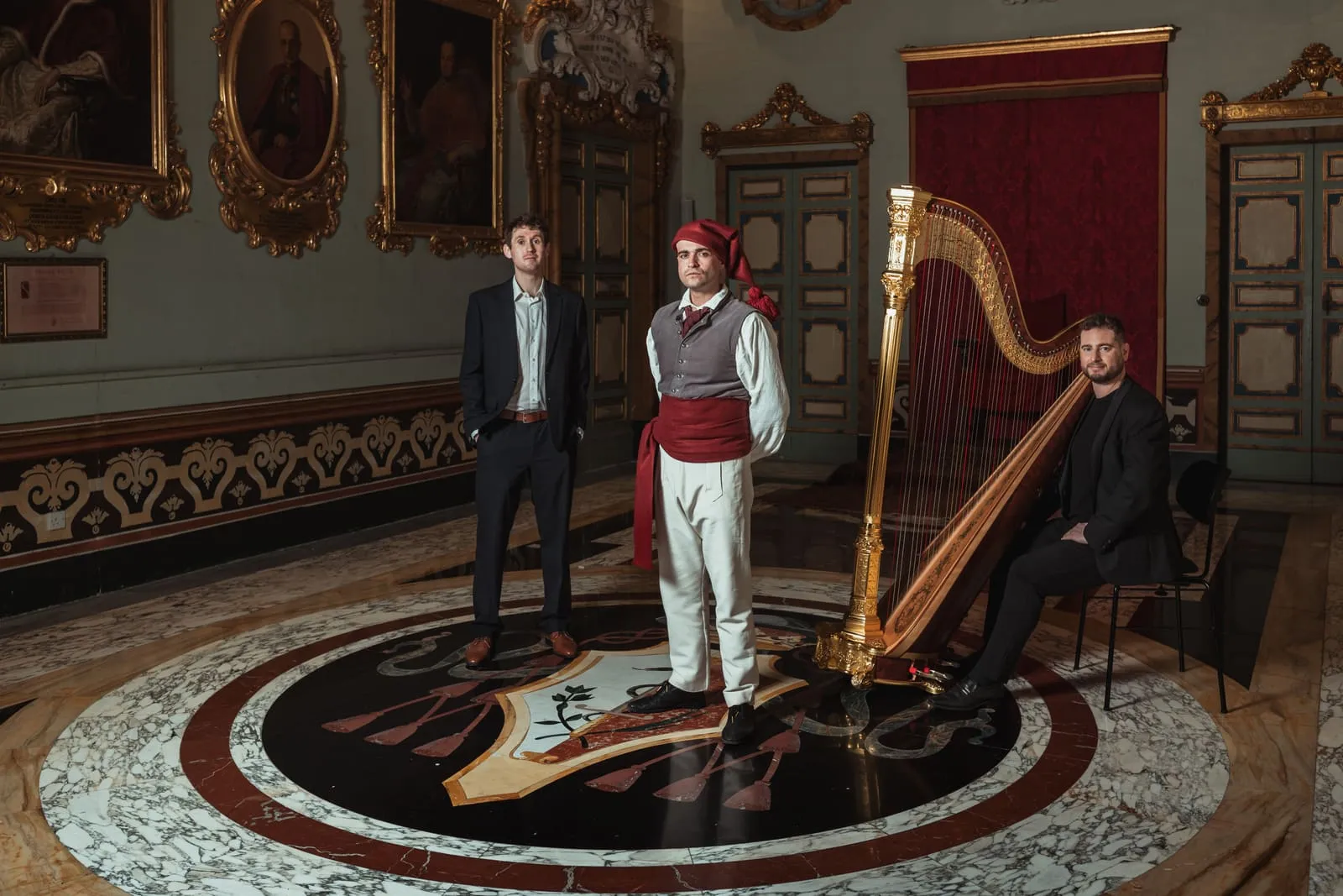 A costumed performer stands between two musicians and a gilded harp on the marble floor of an ornate hall, portraits and red drapes behind them.
