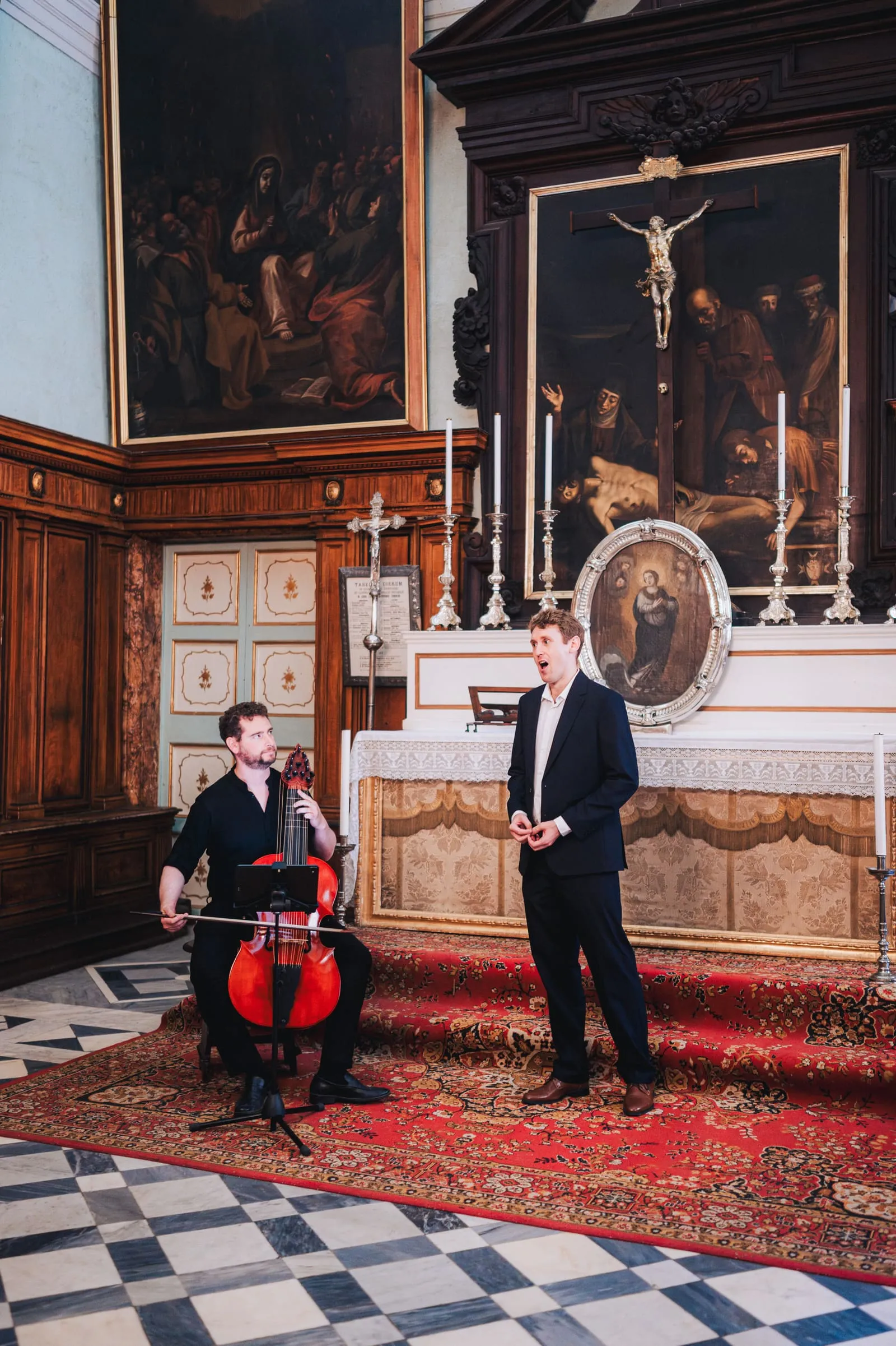 A tenor and a musician on a red Baroque bass viol perform beside an ornate side altar hung with paintings, candlesticks lit around them.