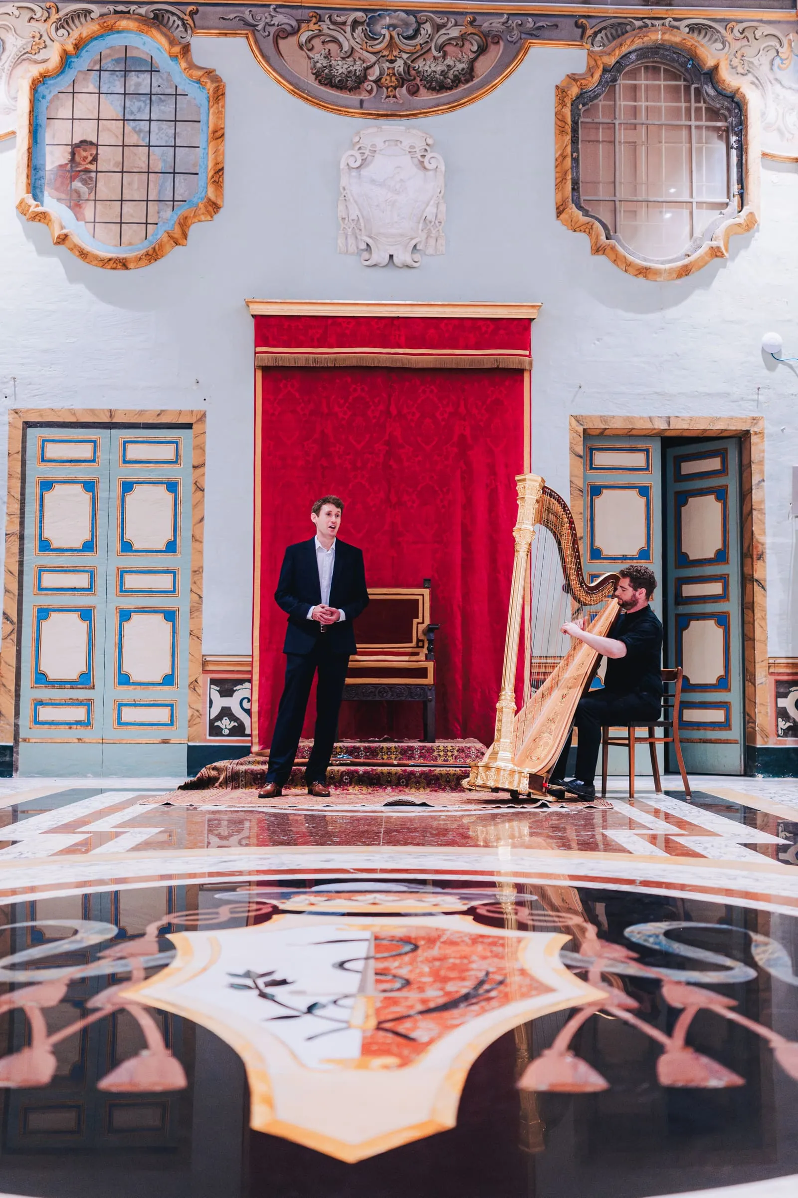 A tenor sings as a harpist plays a gilded harp before a red curtain and throne, a Maltese cross inlaid in the polished marble floor.