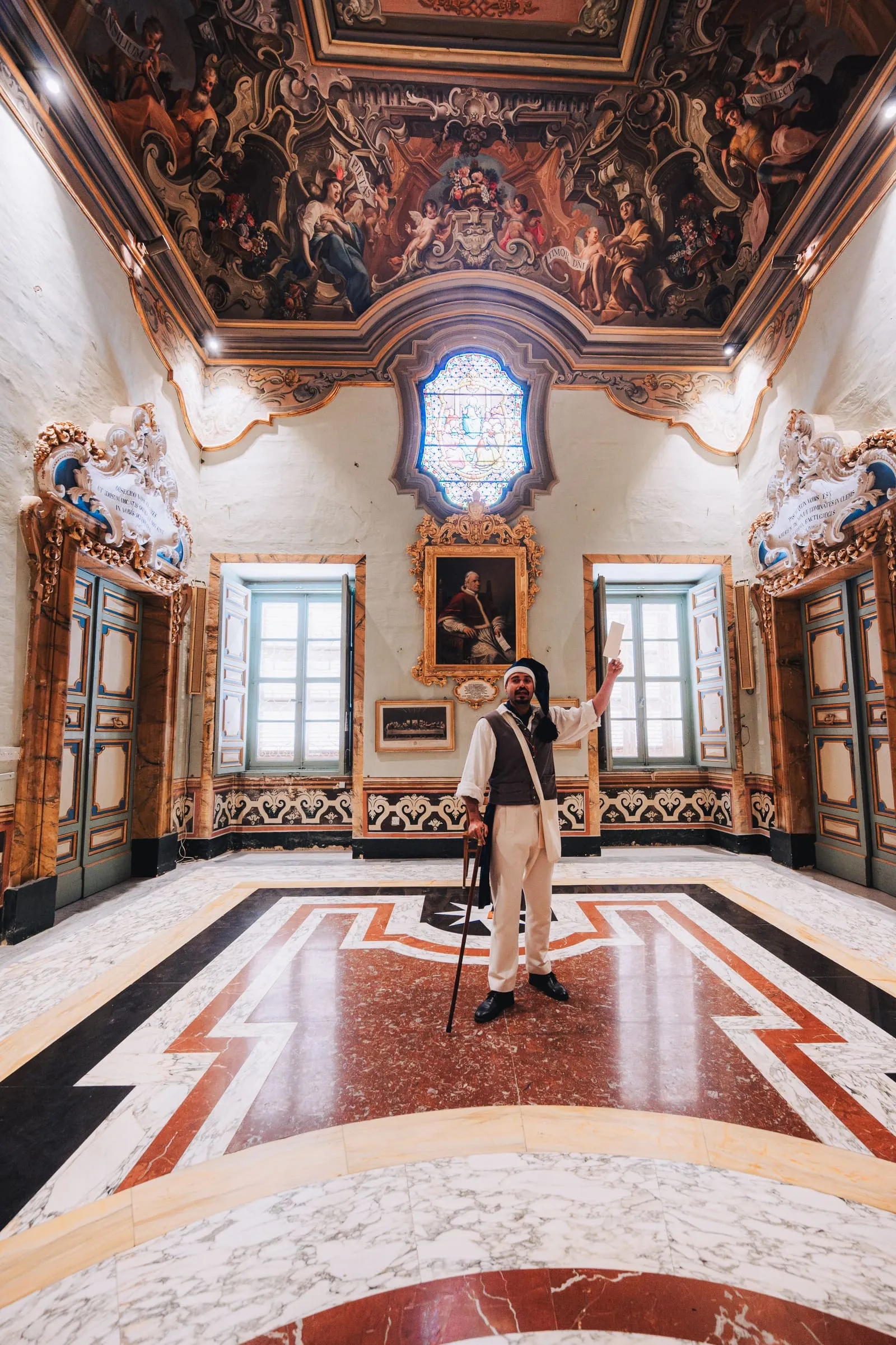 A costumed performer in a nightcap and waistcoat raises a letter aloft in a frescoed marble hall of the Cathedral.