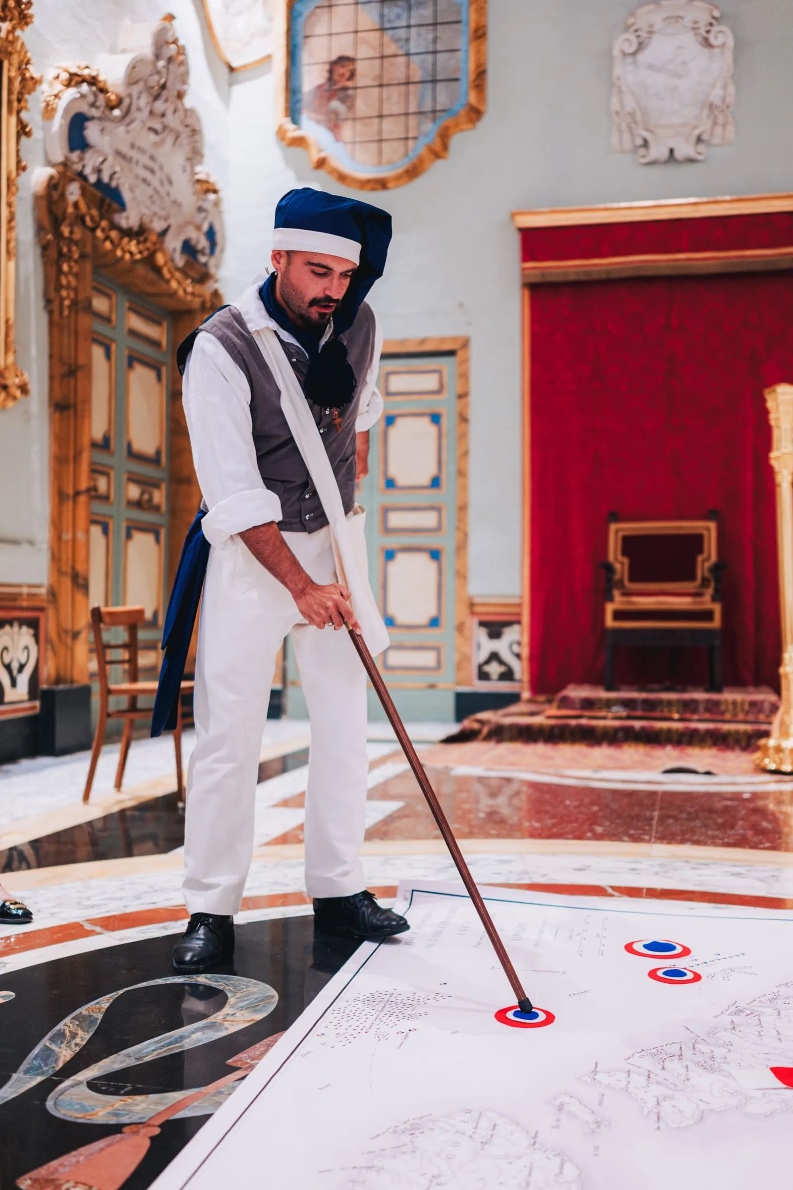 A costumed performer points a cane at a large map of Malta laid across the marble floor, marked with tricolore roundels.