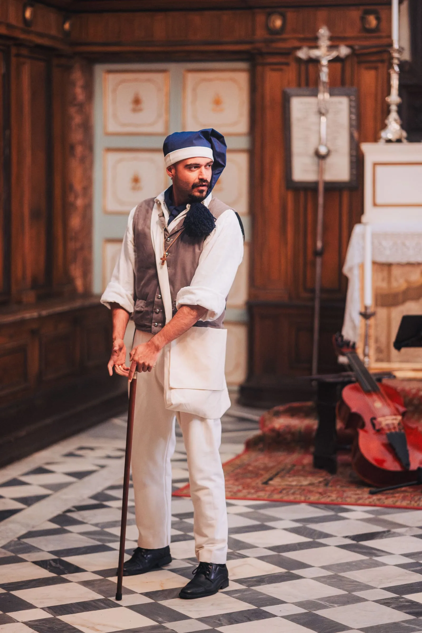 A costumed performer in a blue nightcap leans on a cane on the chequered marble floor, a red Baroque cello resting on a bench behind him.