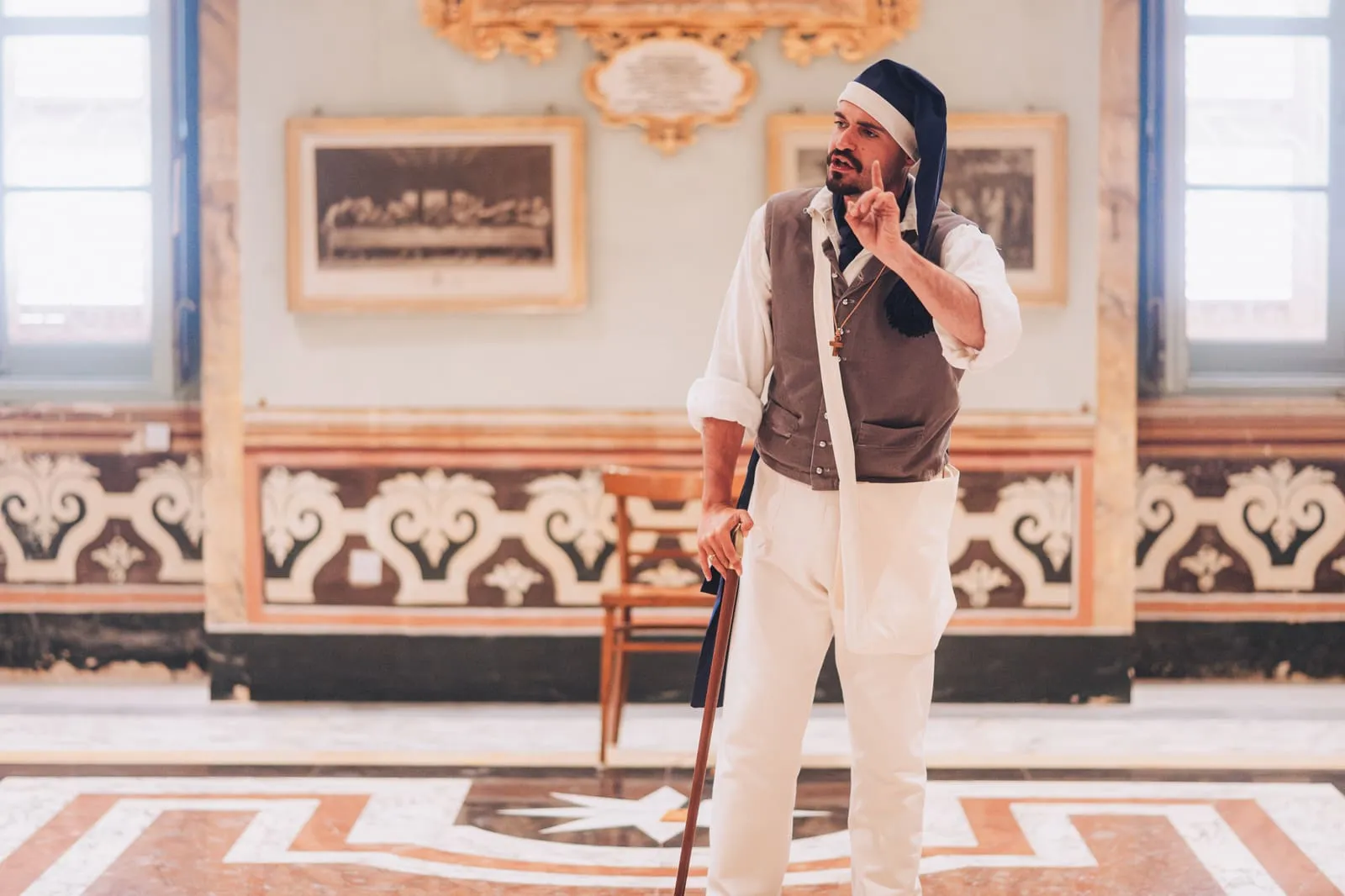 A costumed performer gestures mid-story, finger raised, before an engraving of the Last Supper in a frescoed hall.