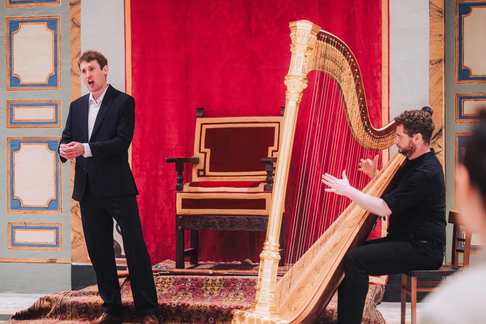 A tenor sings beside a harpist at a gilded harp, framed by a red damask drape and the painted walls of the Cathedral hall.