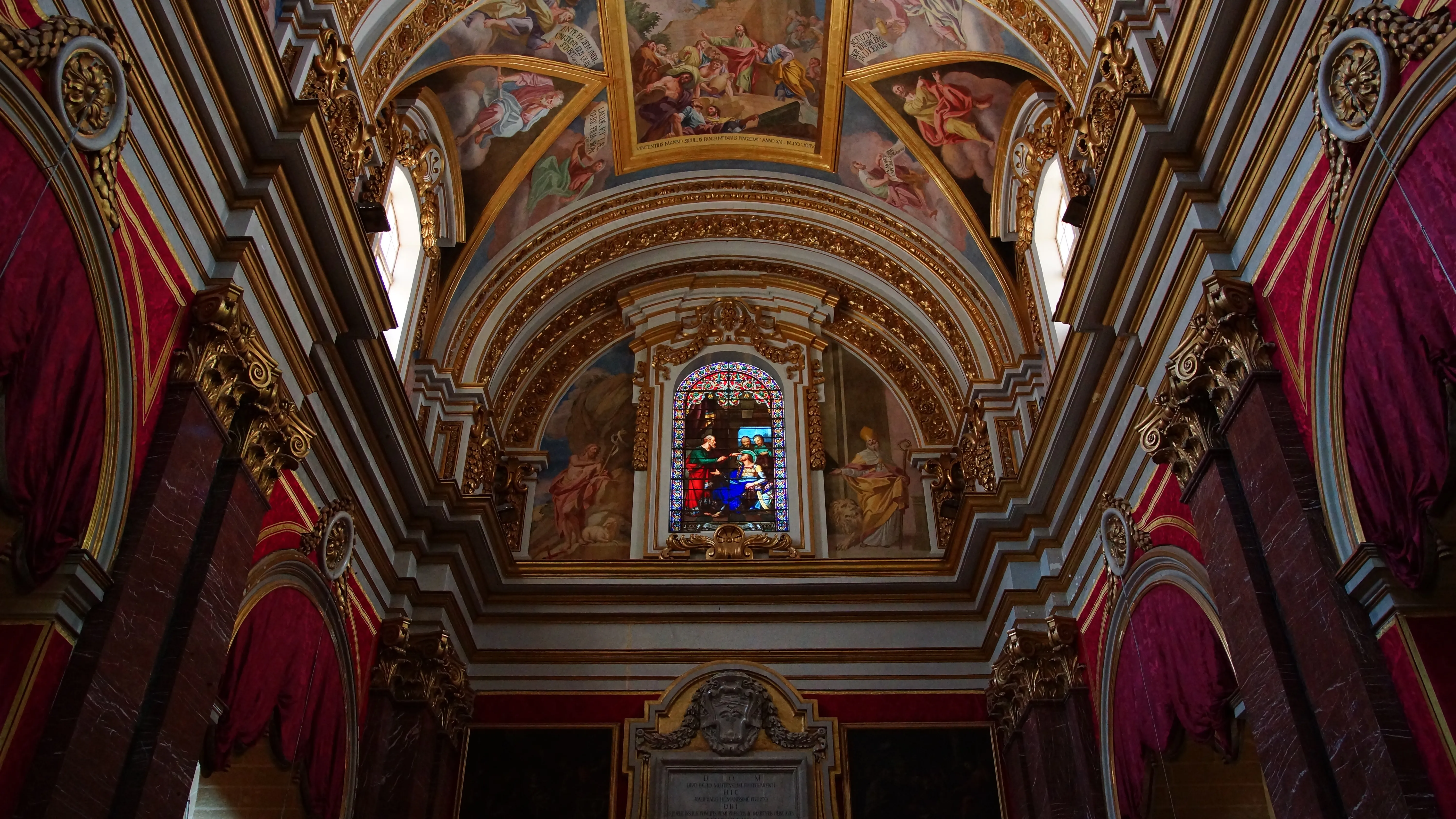 The interior of St Paul's Cathedral, Mdina: a frescoed and gilded vaulted ceiling above a central stained-glass window, framed by marble columns hung with deep-red damask.