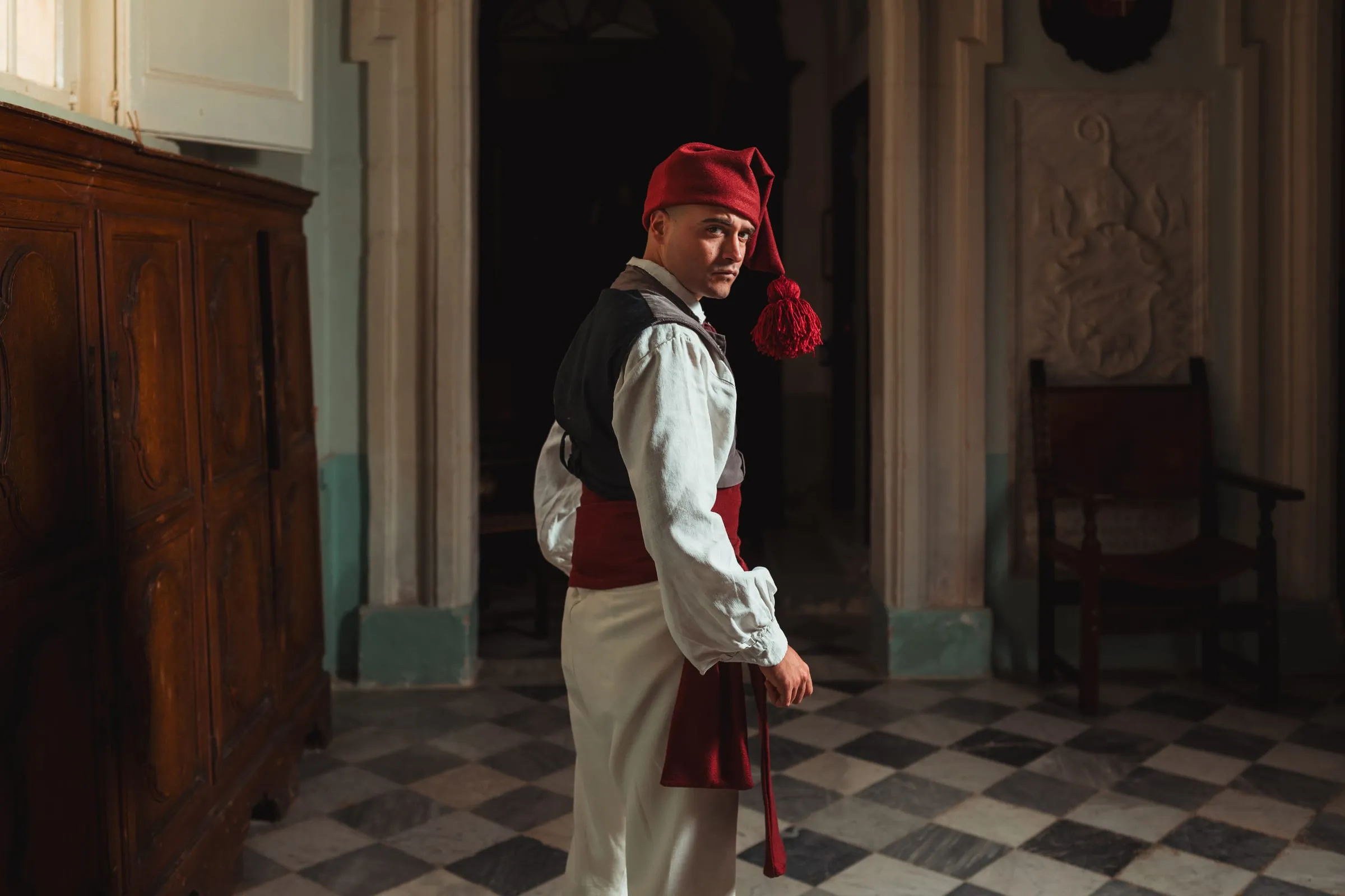 A costumed performer in a red nightcap and sash looks back over his shoulder in a chequered-marble hall of Mdina.