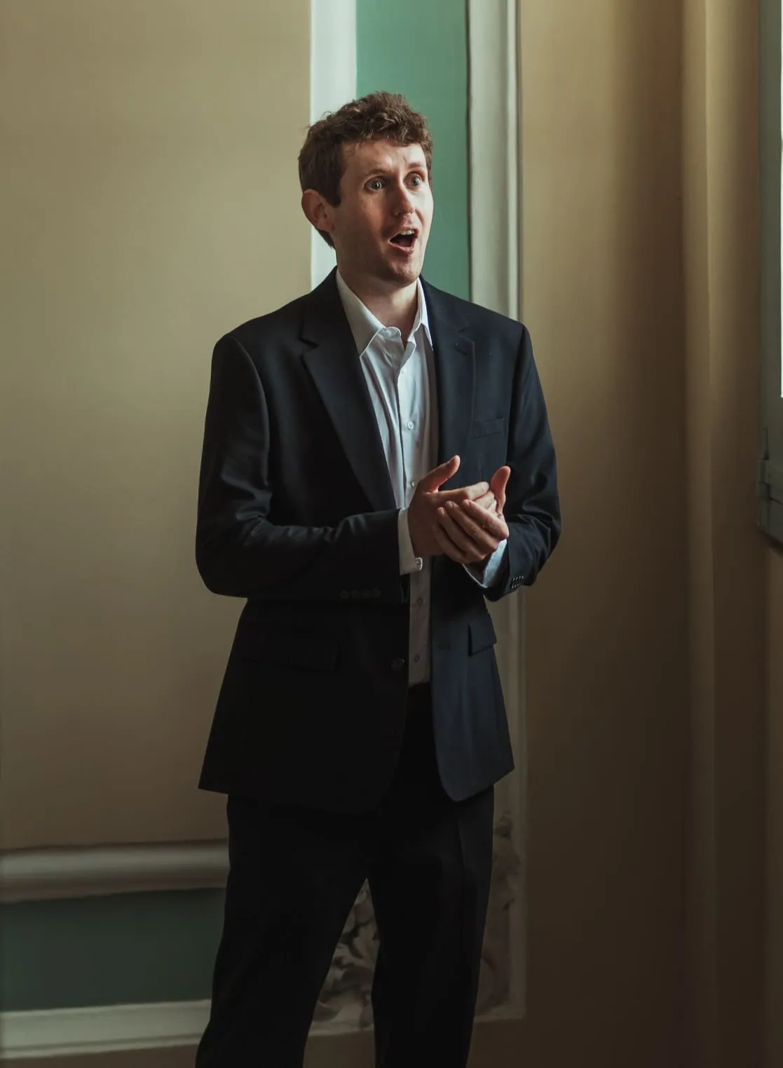 Studio portrait of Tom Armitage, tenor, in a navy suit against a dark background.