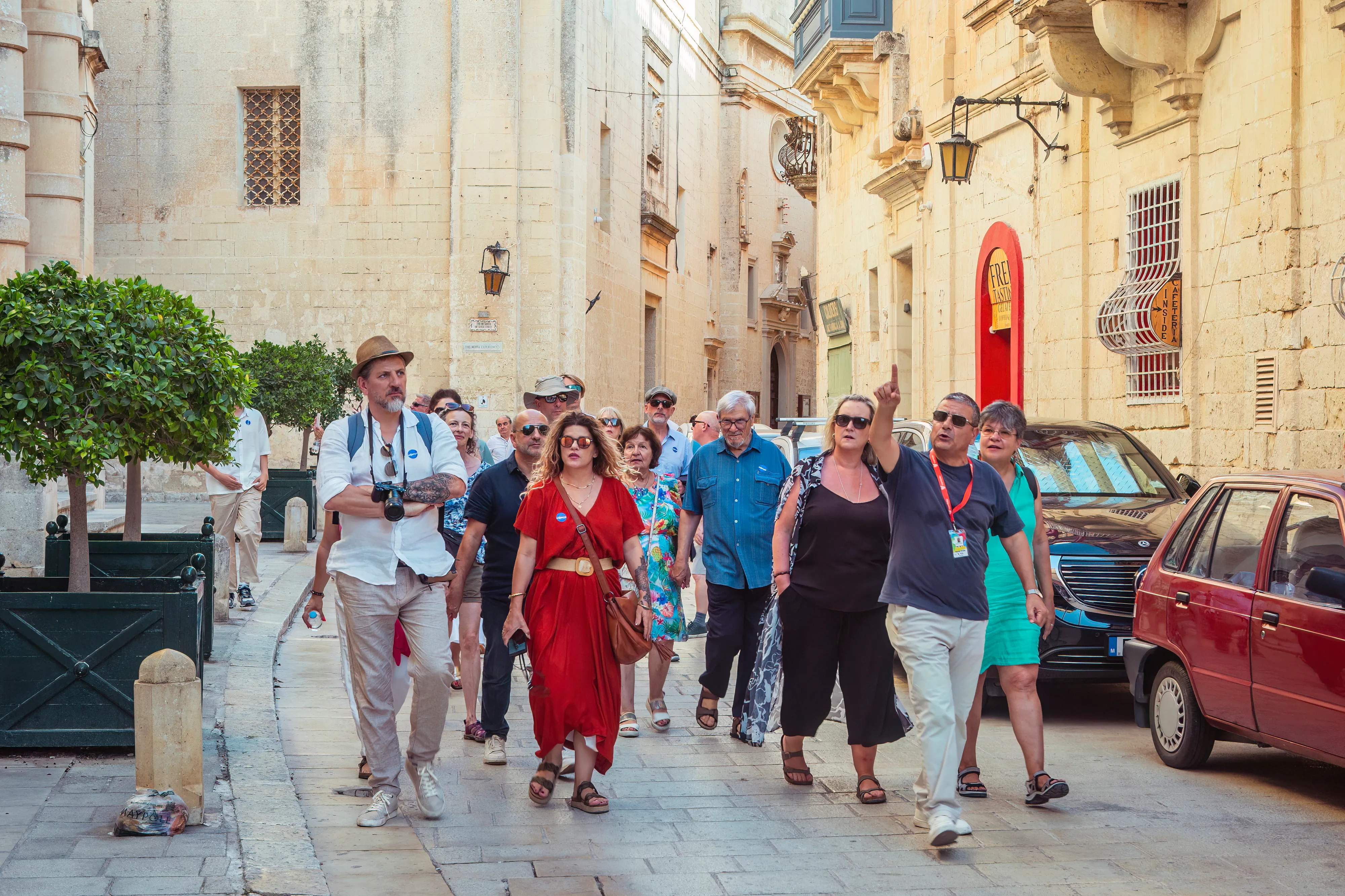 A professional tourist guide leads a group of visitors through a sunlit limestone street in Mdina, pointing up at the historic buildings.